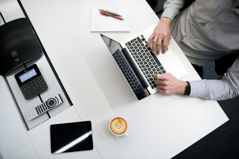 Hybrid Working Man in a shirt working at a white desk, which has a laptop on, a notepad, phone intercom and a coffee